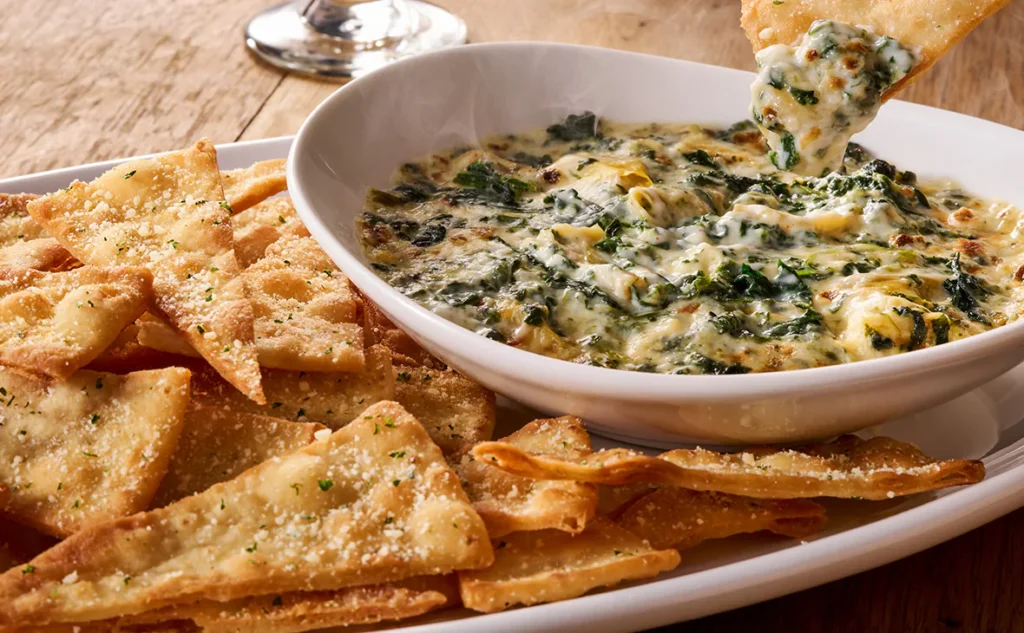 Olive Garden Spinach-Artichoke Dip served warm in a bowl with crispy flatbread chips on the side, topped with melted cheese.